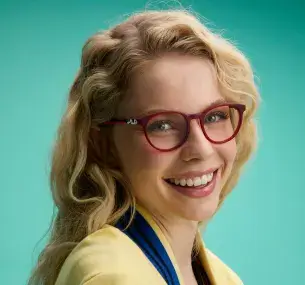 Una donna dalla pelle chiara, con capelli biondi e mossi, è ritratta di profilo mentre sorride. Indossa occhiali da vista rossi con una montatura piccola e leggera, sulle cui aste è stampato il logo FILA.